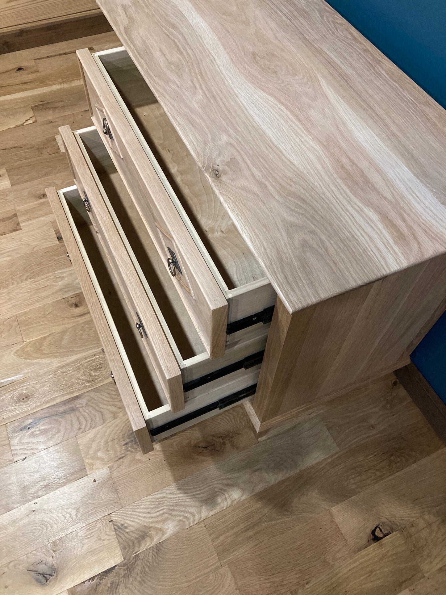 A wooden nightstand with three drawers that are open. The drawers are made of light wood with a natural grain pattern. The dresser is sitting on a light wood floor.