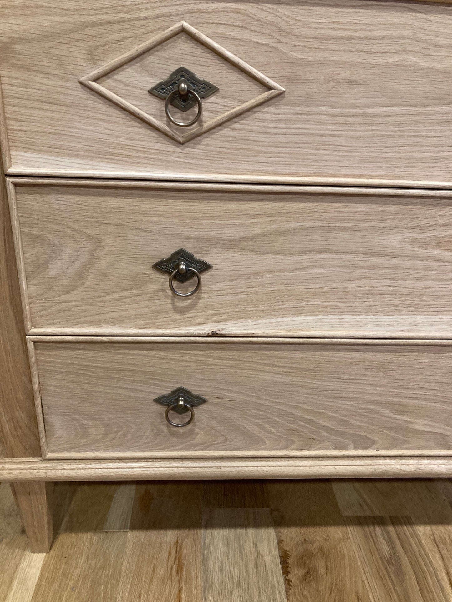 A light wood dresser with three drawers. Each drawer has a decorative metal ring pull. The dresser is sitting on a light wood floor.