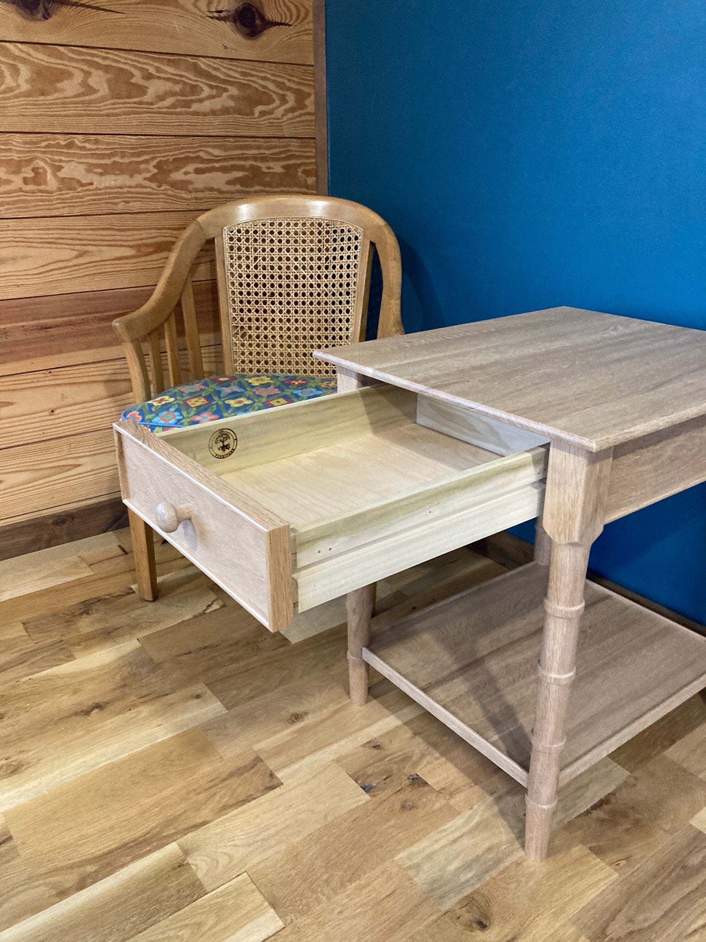 A light wood end table with a drawer pulled out and a lower shelf. The table is next to a wicker chair with a floral patterned cushion.