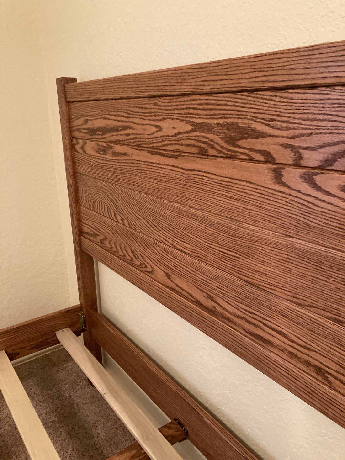A wooden bed frame with a brown stained headboard and a slatted base. The headboard has a simple design with horizontal lines and a slightly curved top.