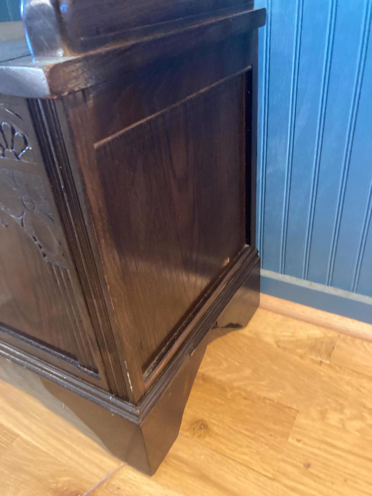 Dark brown wooden storage chest with carved details. The chest features a rectangular shape with a slightly tapered base. The wood grain is visible, and there are subtle carvings on the front panel. The chest sits against a blue wall and light brown wood floor. Antique style wooden chest, vintage storage trunk, dark wood furniture.