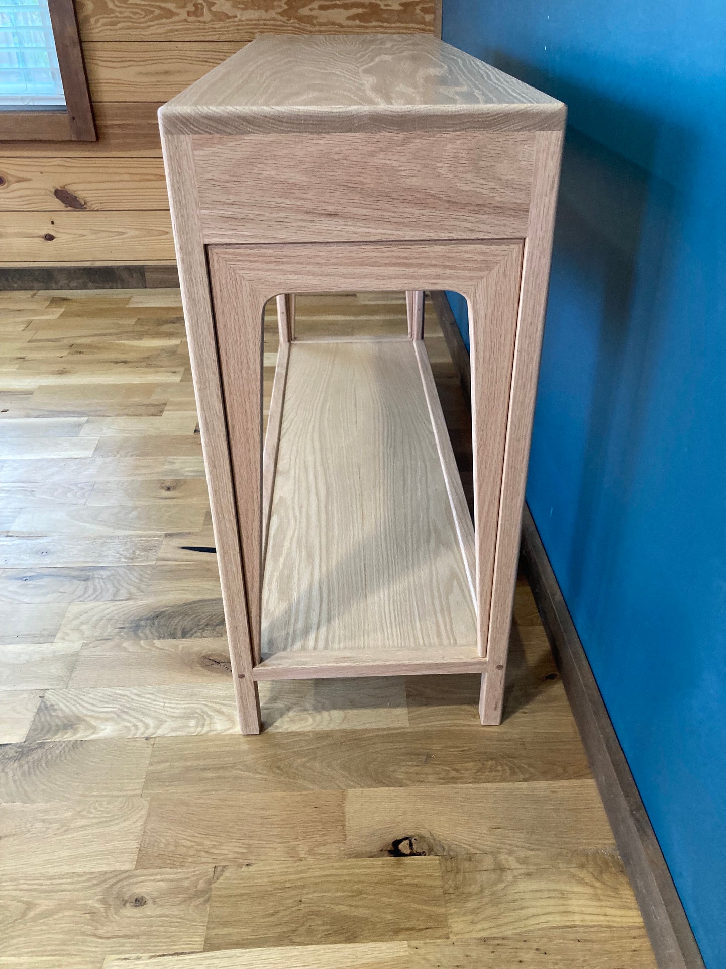 A side view of a light wood console table with a bottom lower shelf and drawers. The table is standing on a light wood floor with a blue wall in the background.