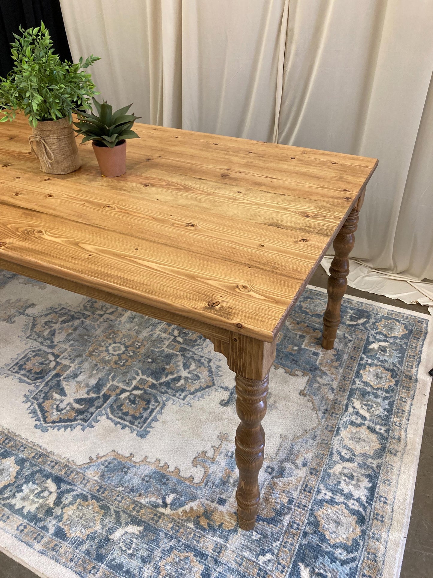 A wooden dining table with a light brown finish and turned legs. The table is on a rug with a blue and white pattern.