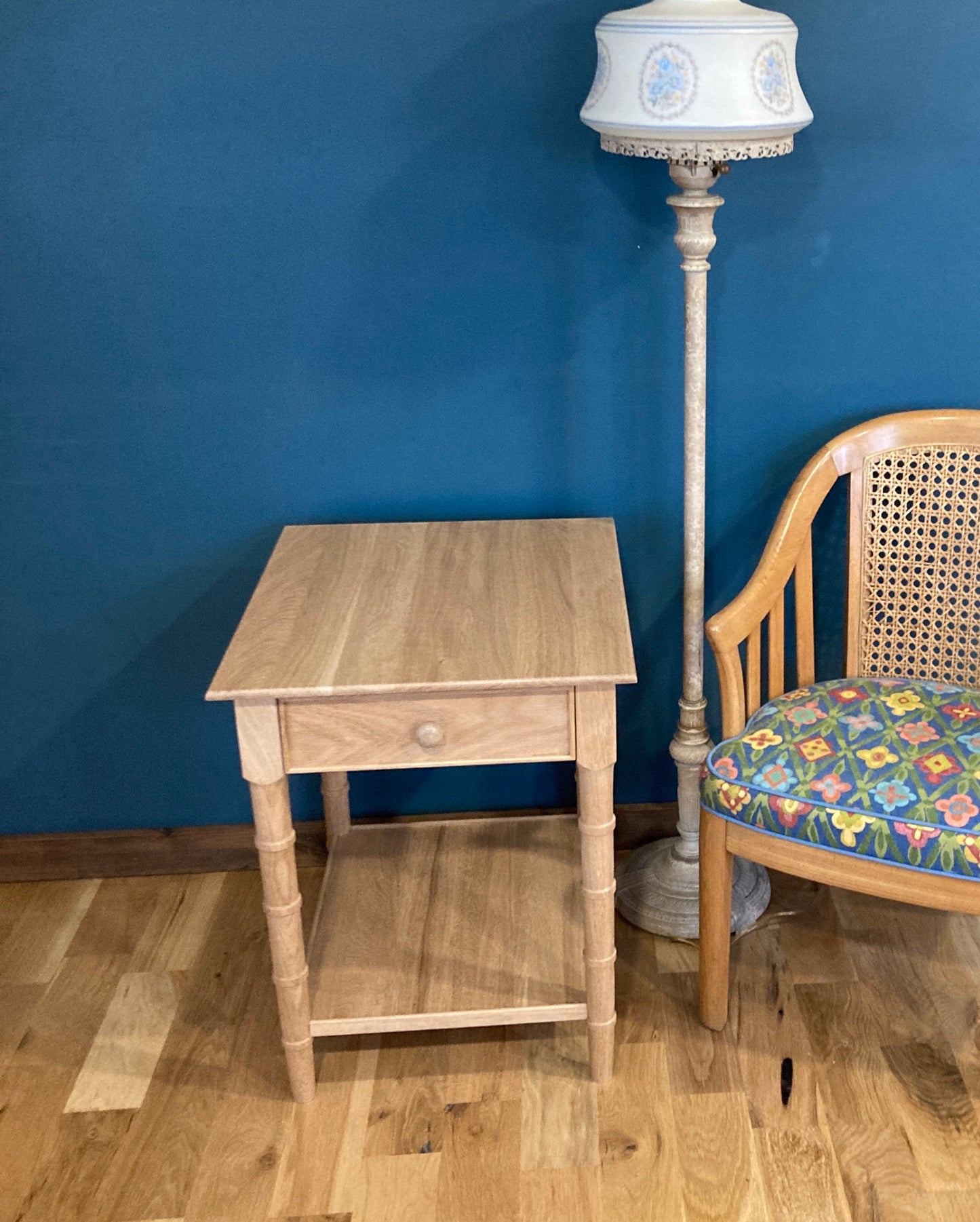 A light wood end table with a single drawer and a lower shelf. The table has a simple design with turned legs. The table is in front of a blue wall and next to a chair with a floral patterned cushion.