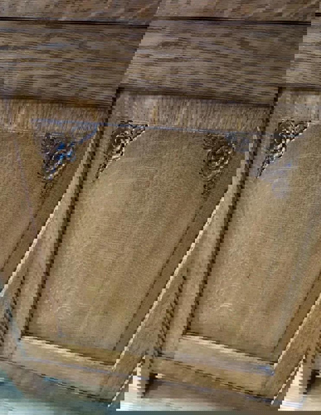 A close-up of a wooden cabinet door with two carved floral designs in the top corners. The door is made of light brown wood with a natural wood grain pattern.