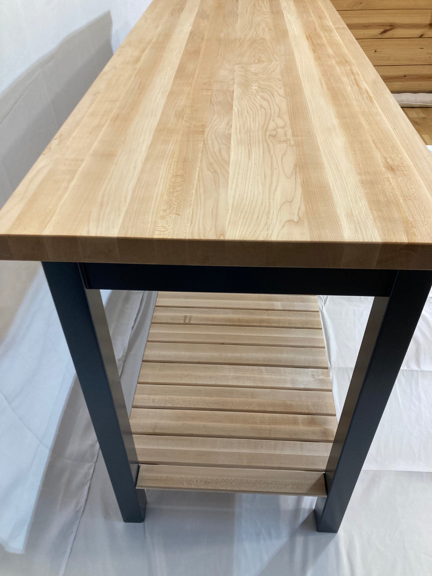 A kitchen island with a light maple butcher block countertop and a blue painted frame. The island features a lower shelf made of light maple wood slats. The island's clean lines and simple design create a modern aesthetic, suitable for various kitchen styles. The butcher block top is durable and easy to maintain, while the blue legs provide a sturdy and stylish base. The open shelf offers convenient storage space.
