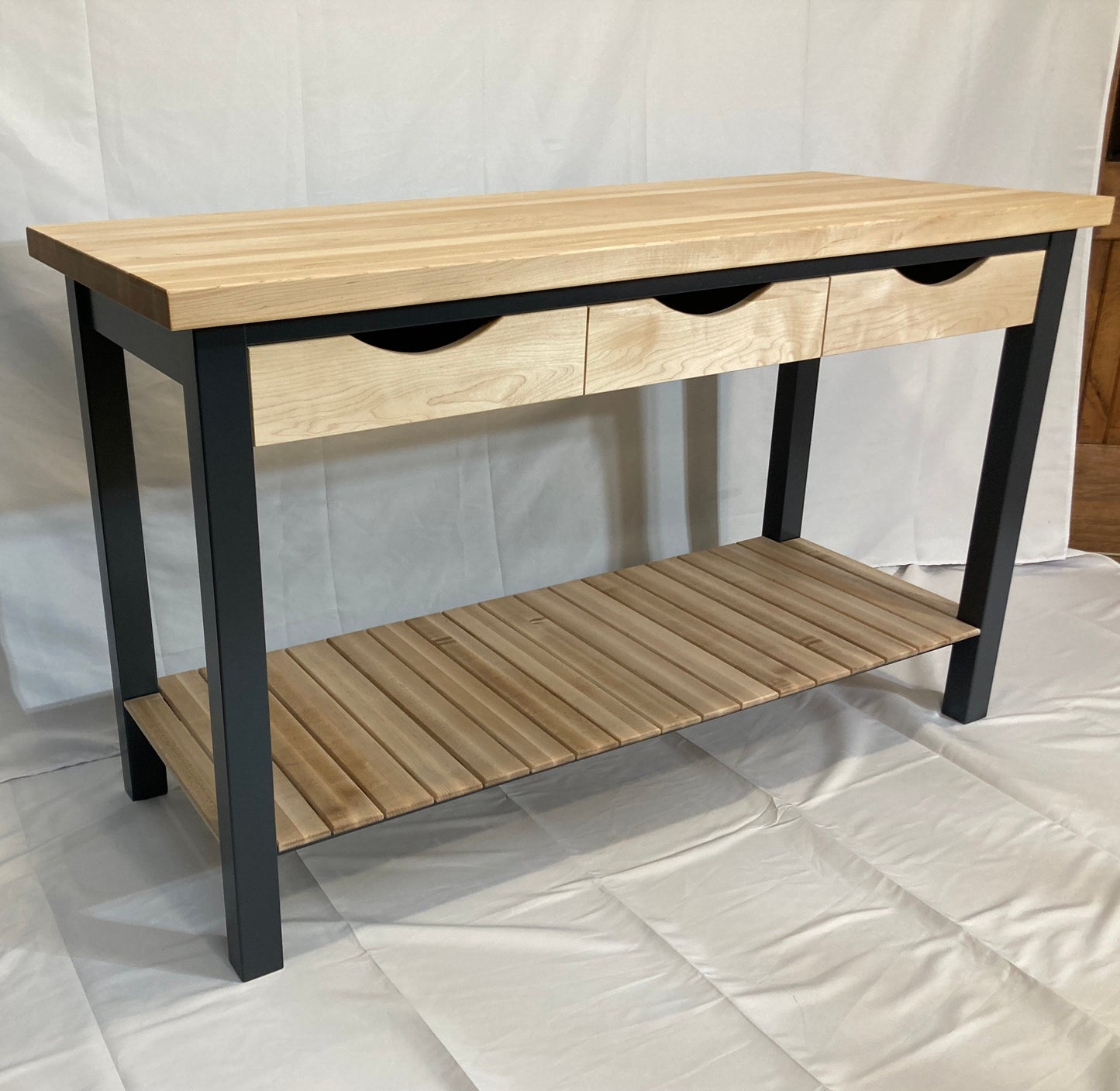 A kitchen island with a light maple butcher block top and three drawers. The island features a lower shelf with slatted wood and a blue painted wood frame. The drawers have a curved, recessed design. This kitchen island is ideal for adding extra counter space and storage to a kitchen.