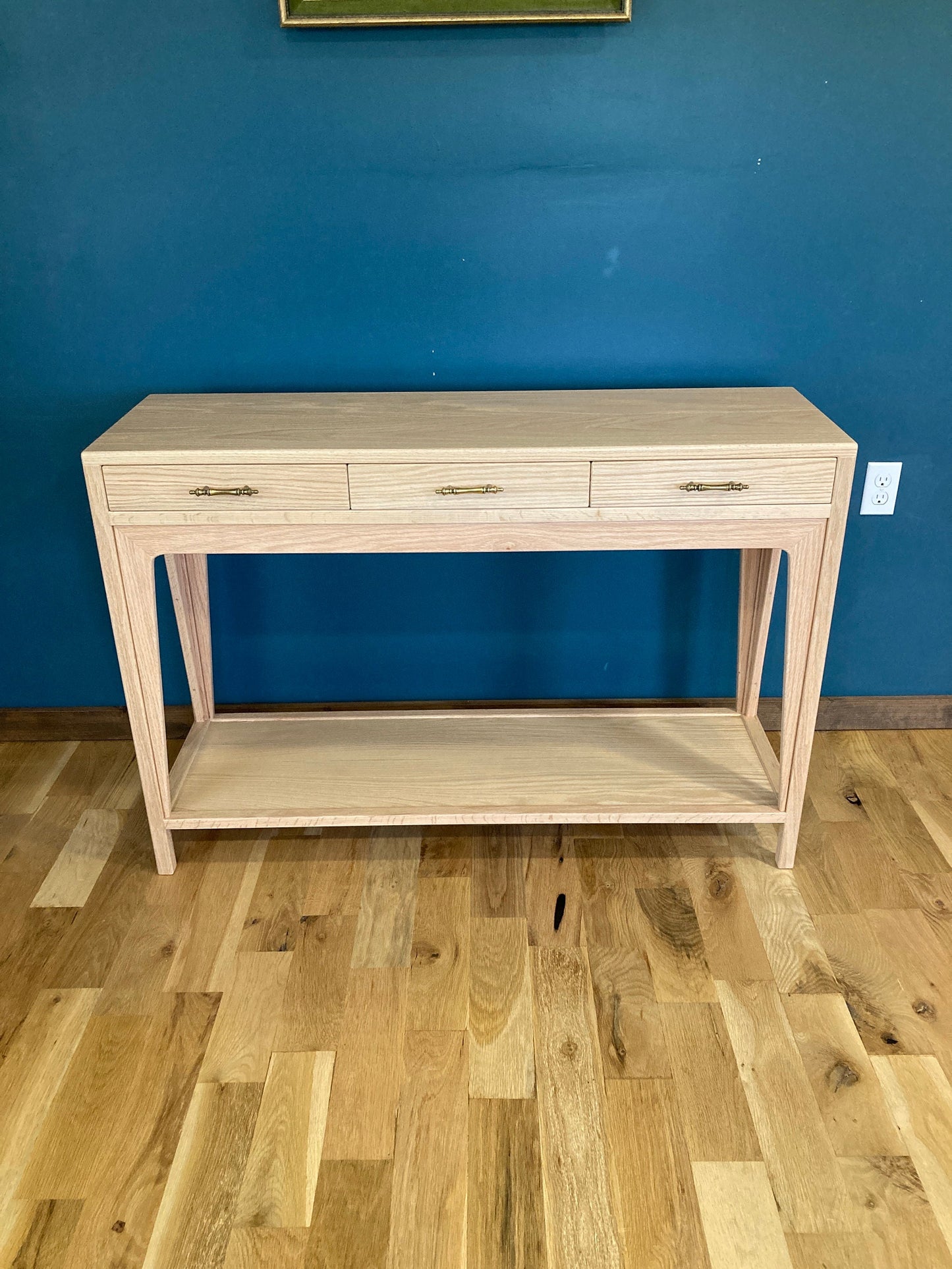 A light wood console table with three drawers and a lower shelf. The drawers have brass pulls. The table is standing on a light wood floor.