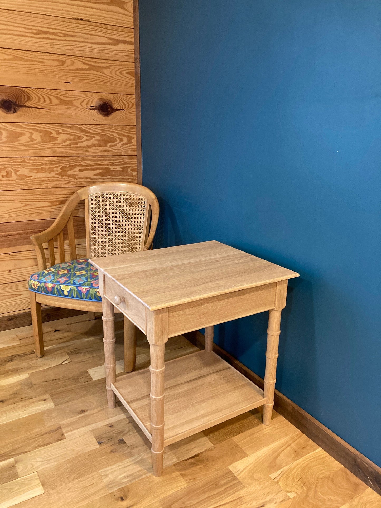 A light brown wooden end table with a drawer and a lower shelf. The table is in a room with light brown wood floors and a blue wall. A chair with a floral patterned cushion is next to the table.