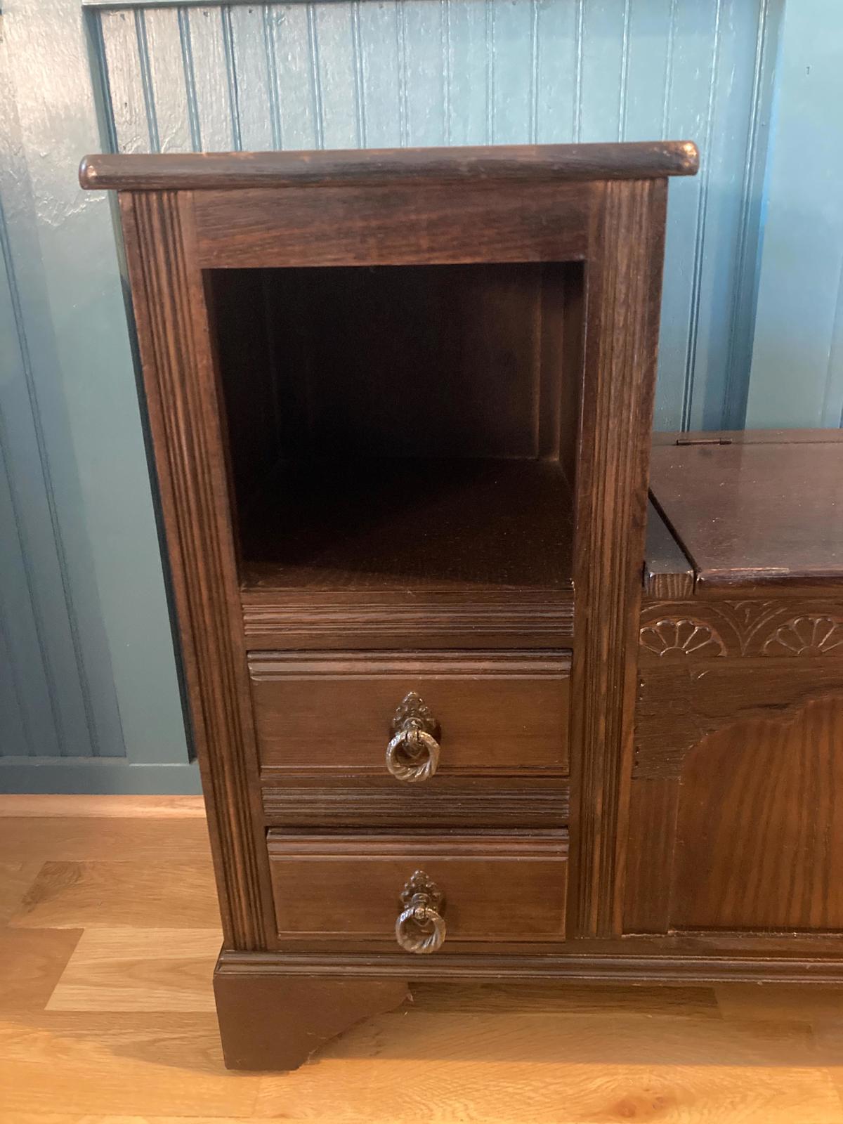 A dark brown wooden storage cabinet with two drawers and an open shelf. The drawers have antique-style ring pulls. The cabinet has a paneled door with carved details. The piece is an antique, made of oak. The cabinet is free-standing and sits on a light brown wood floor against a teal wall.