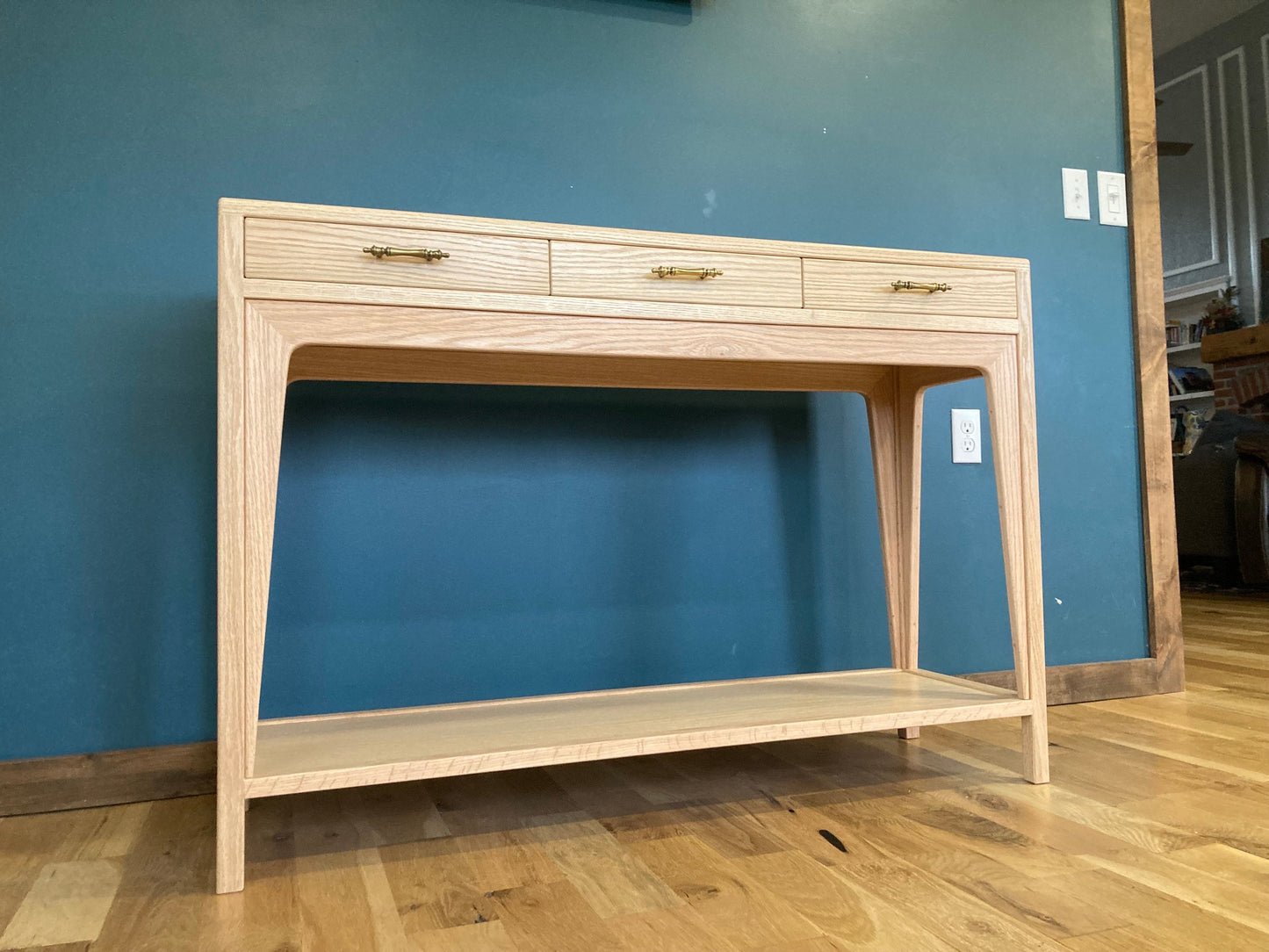 A light wood console table with three drawers and a lower shelf. The drawers have brass pulls. The table is standing on a wood floor.