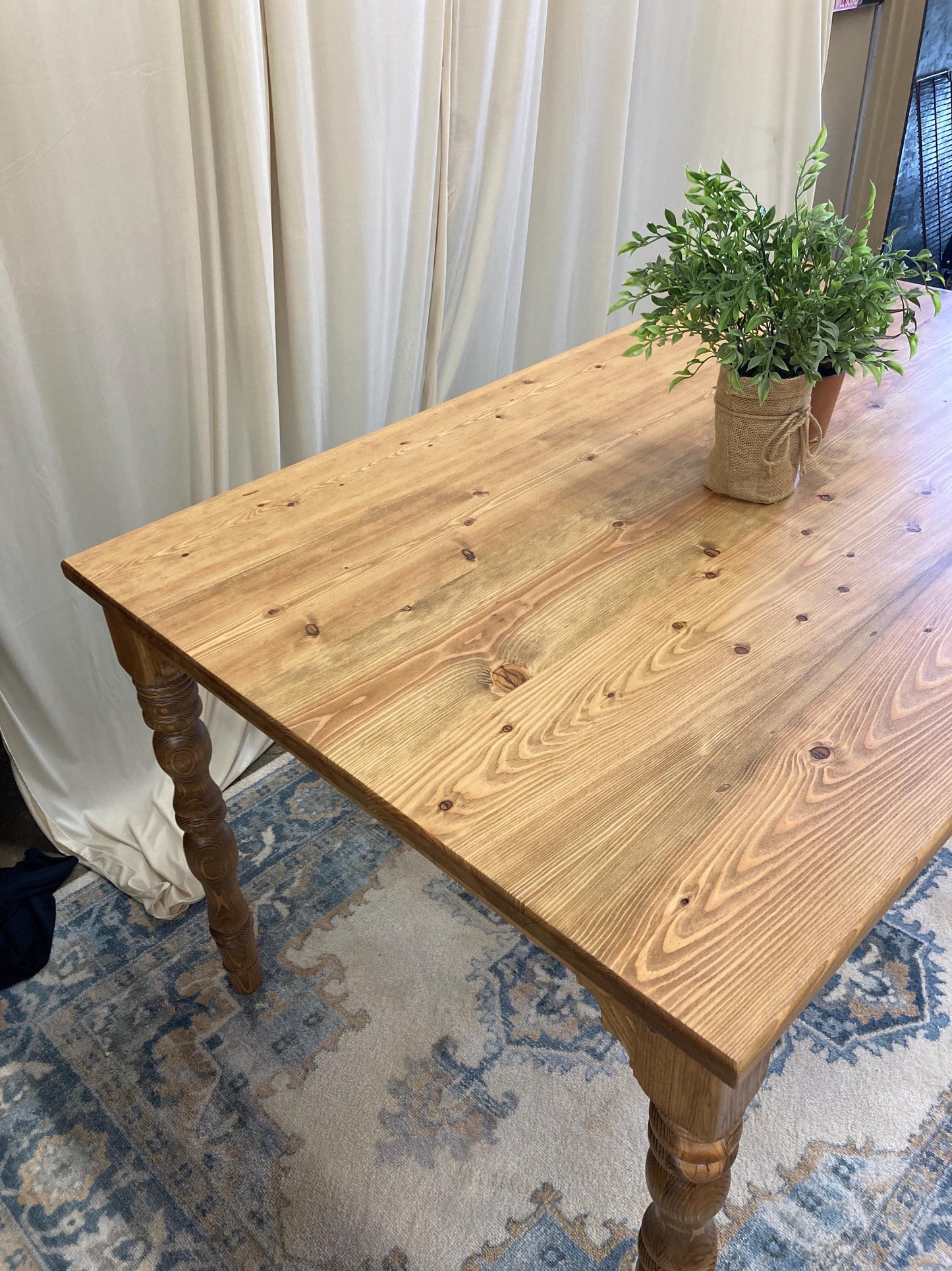 A wooden table with a light brown finish and turned legs. The table is on a rug with a blue and beige pattern.