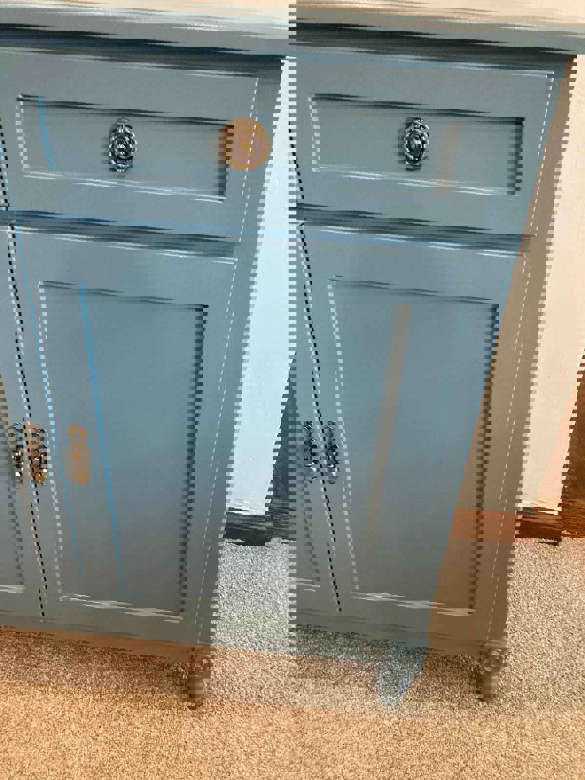 close up view of a blue cabinet drawer and door. The cabinet has brass hardware and sits on four legs. The cabinet is on a light brown carpet.