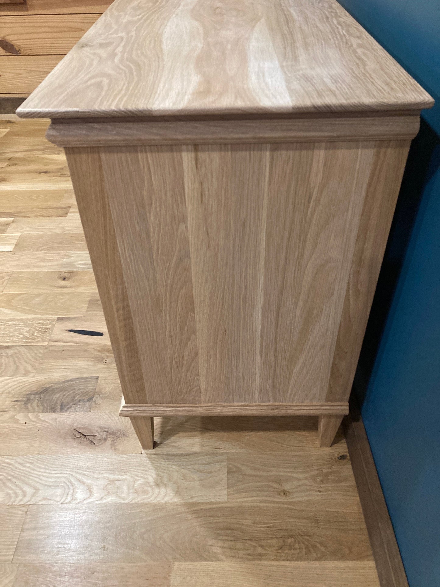 A light wood nightstand with a simple design. The nightstand has a smooth, flat top and a rectangular body with a slightly raised base. The nightstand is made of light wood with a natural grain pattern. The nightstand is standing on a light wood floor.
