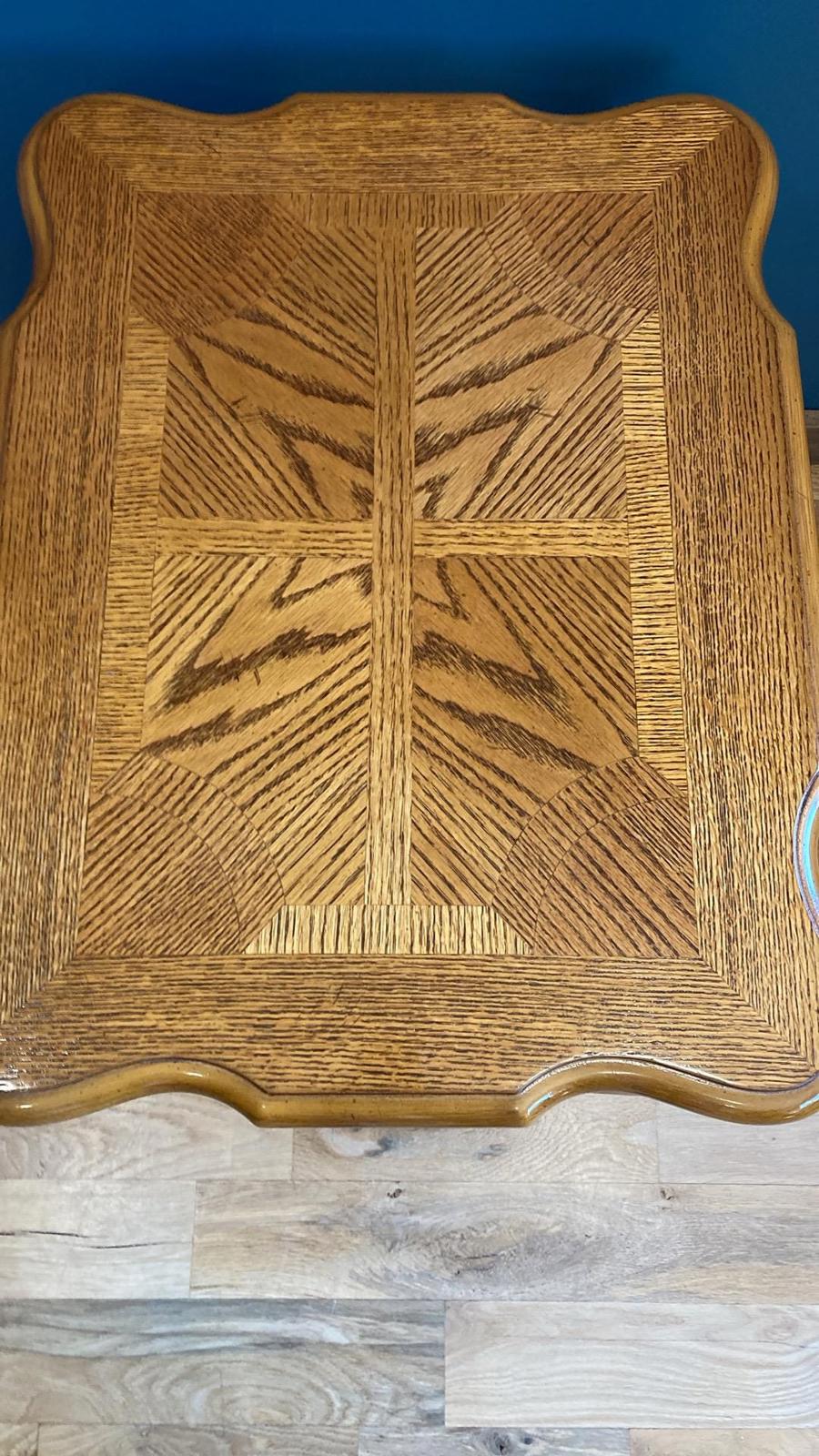 A wooden coffee table with a carved, geometric inlay. The table top is made of light brown oak wood with a rich, warm tone. The inlay features a repeating pattern of lines and shapes, creating a visually appealing design. The edges of the table top are slightly curved, adding to its unique aesthetic. The table's overall style is classic and elegant, suitable for a traditional or transitional interior design setting.