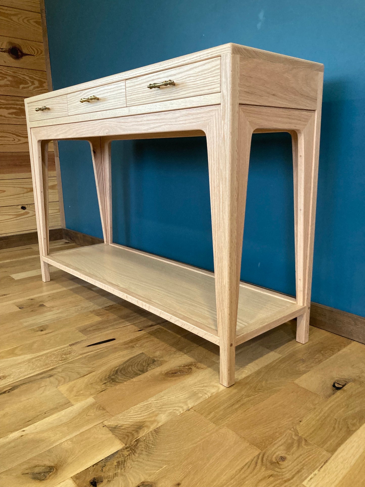 A light wood console table with three drawers and a lower shelf. The drawers have brass pulls. The table is standing on a light wood floor.
