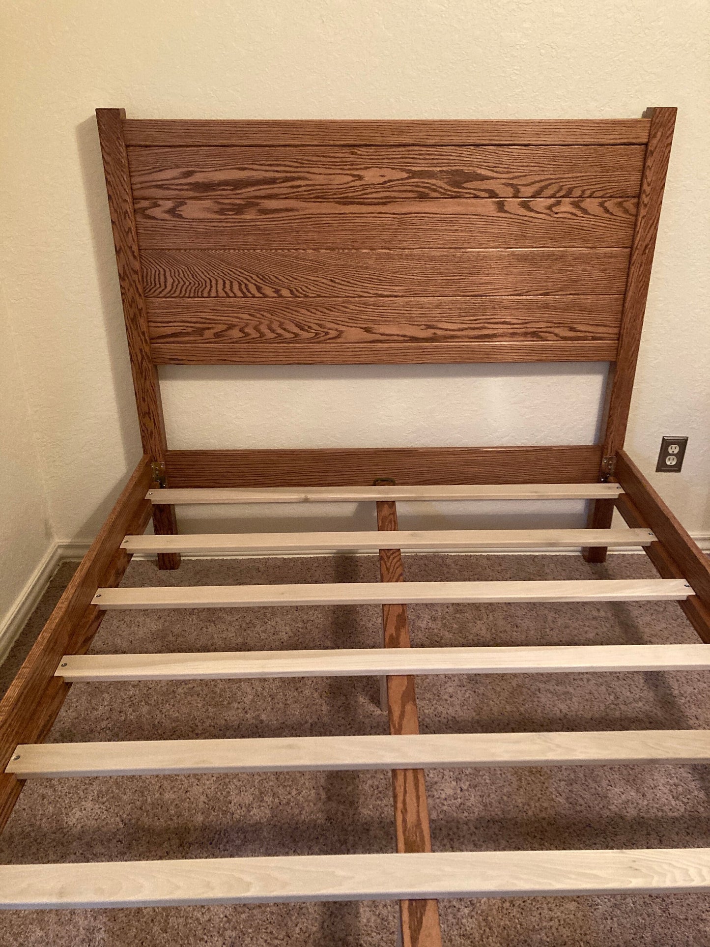 A wooden bed frame with a brown headboard and a light brown slatted base. The bed frame is sitting on a brown carpet.