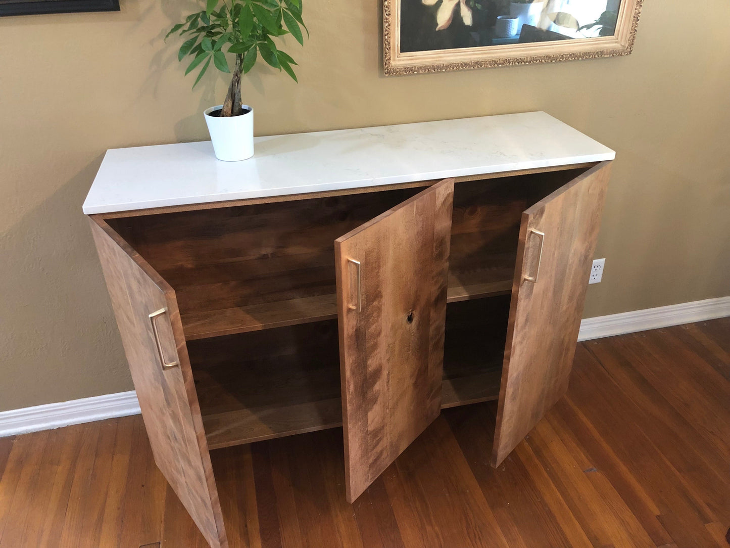 A wooden cabinet with a white quartz stone top and three doors. The doors are open, revealing two shelves inside. The cabinet is made of a light brown wood with a natural wood grain finish. The cabinet has gold-colored handles.