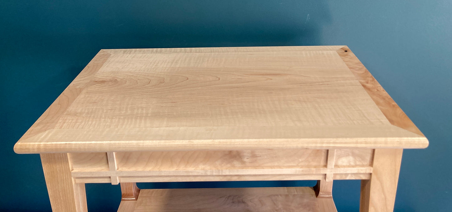 A light-colored wooden end table with a minimalist design. The table features a rectangular top, a lower shelf, and clean lines. The wood grain is visible, showcasing a light, natural finish. The piece is crafted from solid wood, adding to its durability and aesthetic appeal. This simple yet elegant side table would complement various home decor styles.