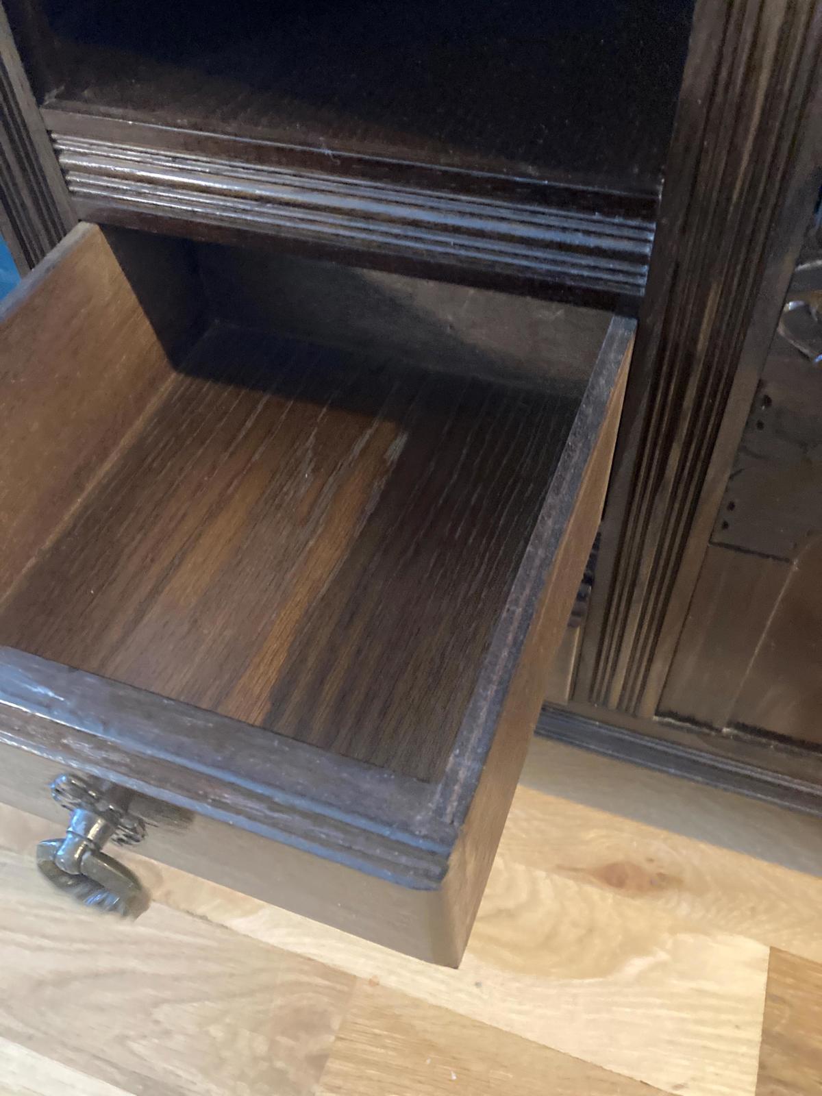 Close-up view of a dark brown wooden drawer pulled open, revealing the interior. The drawer features a simple, ornate metal pull. The wood grain is visible, showing a dark, rich tone. The drawer sits within a dark brown wooden chest, with visible shelving above. The wood appears to be stained. The overall style suggests antique or vintage furniture.