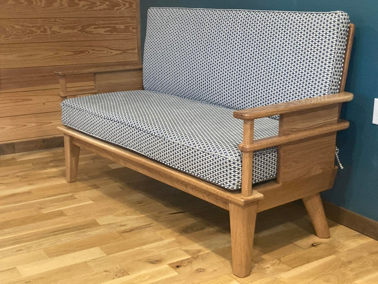 Handmade oak couch with a modern open-arm frame design, upholstered in blue and white houndstooth-style fabric, positioned on a wood floor next teal wall. The couch has wood arms and midcentury style legs.