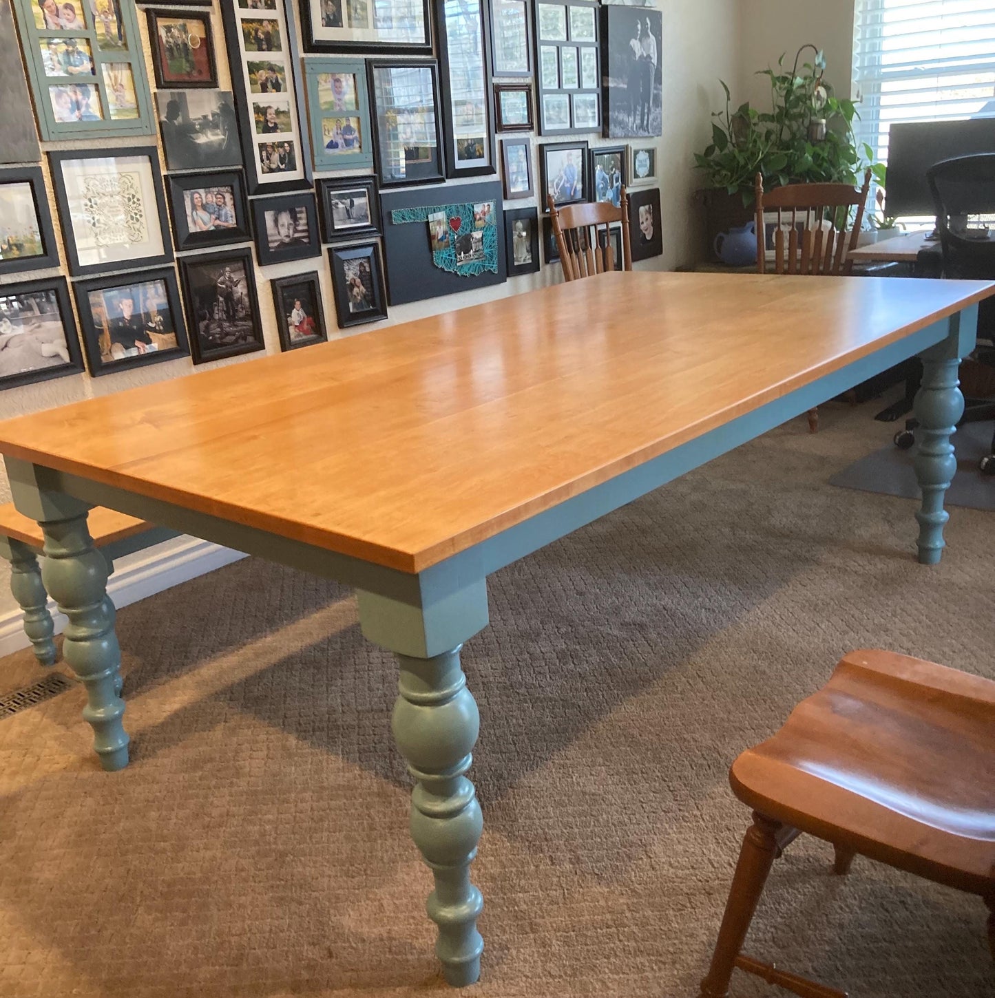 A light wood table with a blue painted base and hand turned legs. The table is in a room with a carpeted floor and a wall with framed pictures.