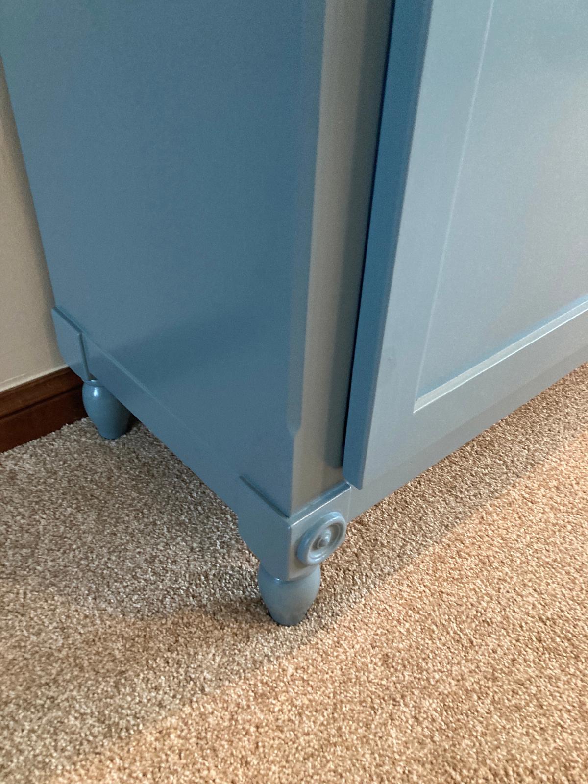 A close up view of the corner of a light blue cabinet edge and hand turned leg. The cabinet is sitting on a beige carpet.
