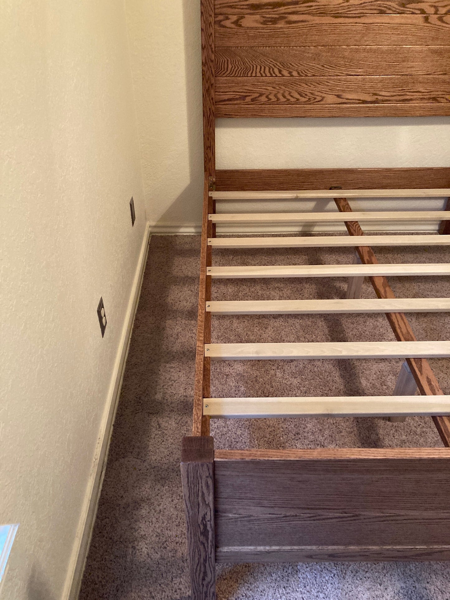 A wooden bed frame with a brown stain. The bed frame has a slatted headboard and a slatted footboard. The bed frame is sitting on a light brown carpet.