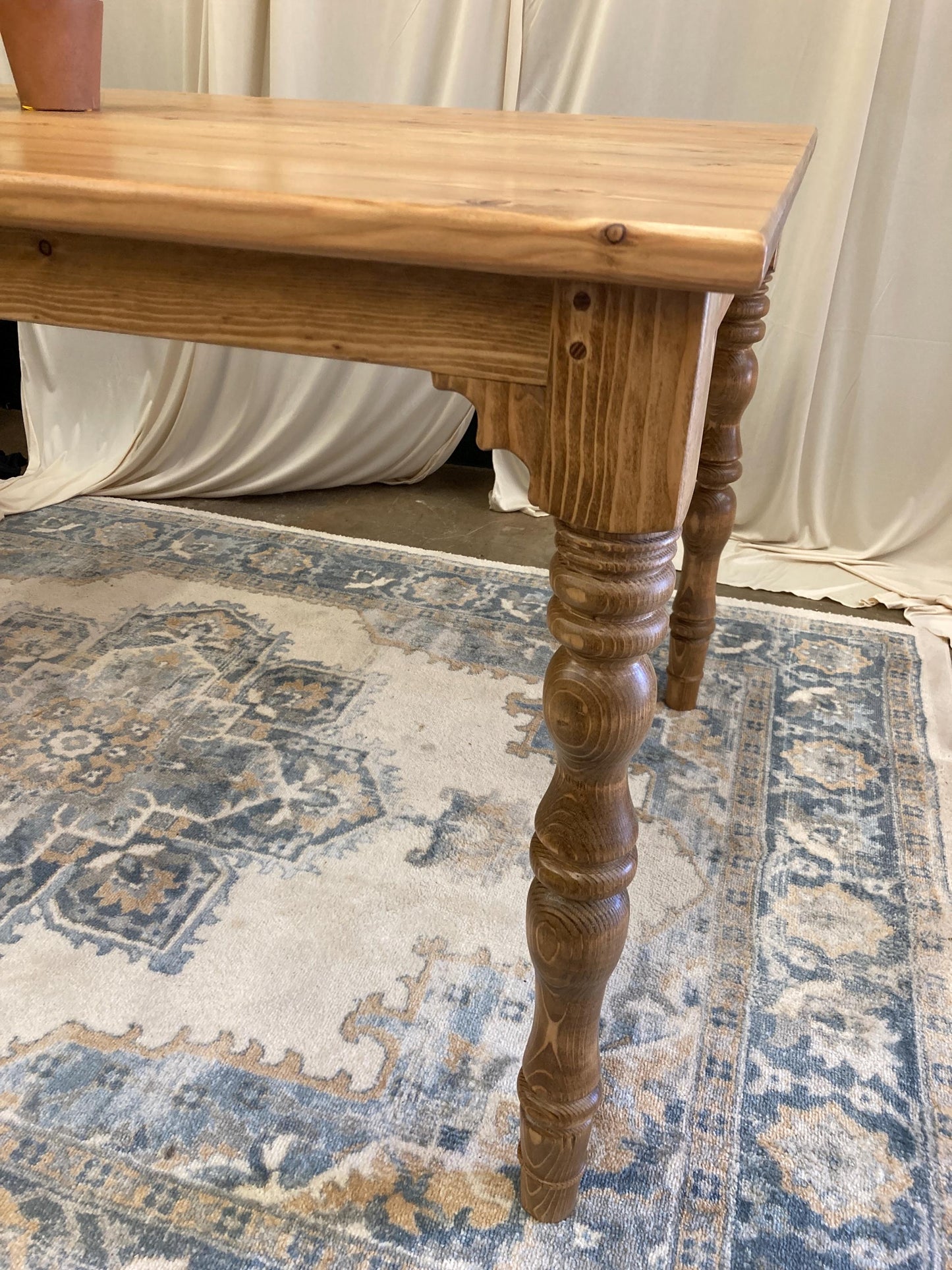 A wooden table with a light brown finish. The table has a rectangular top and four turned legs. The table is sitting on a rug with a blue and brown pattern.