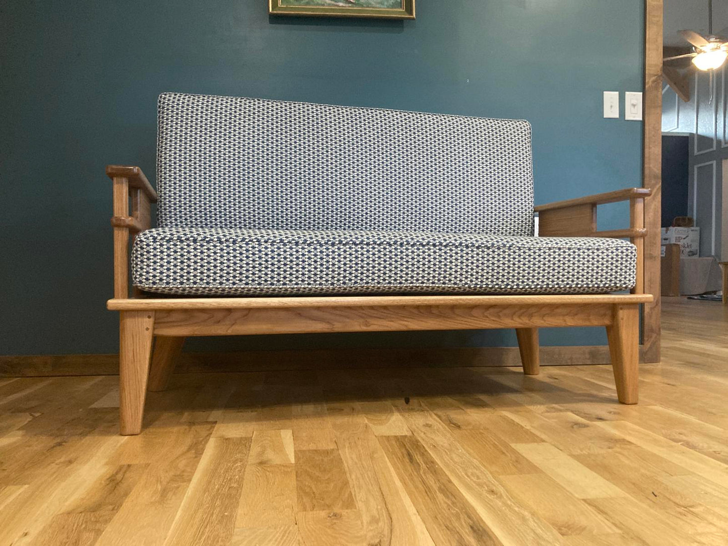 Front view of a solid wood couch with upholstered cushions. It is sitting on a wood floor in front of a teal wall.