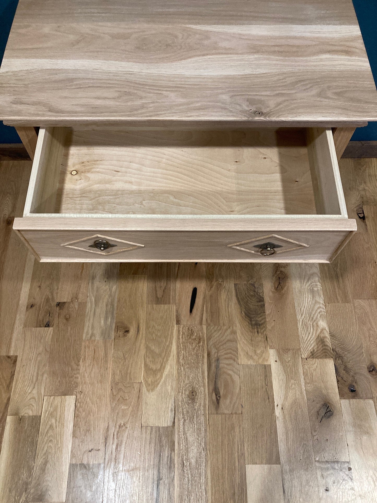 A light wood drawer with a diamond-shaped design on the front. The drawer is open and empty. The drawer is on a light wood floor.