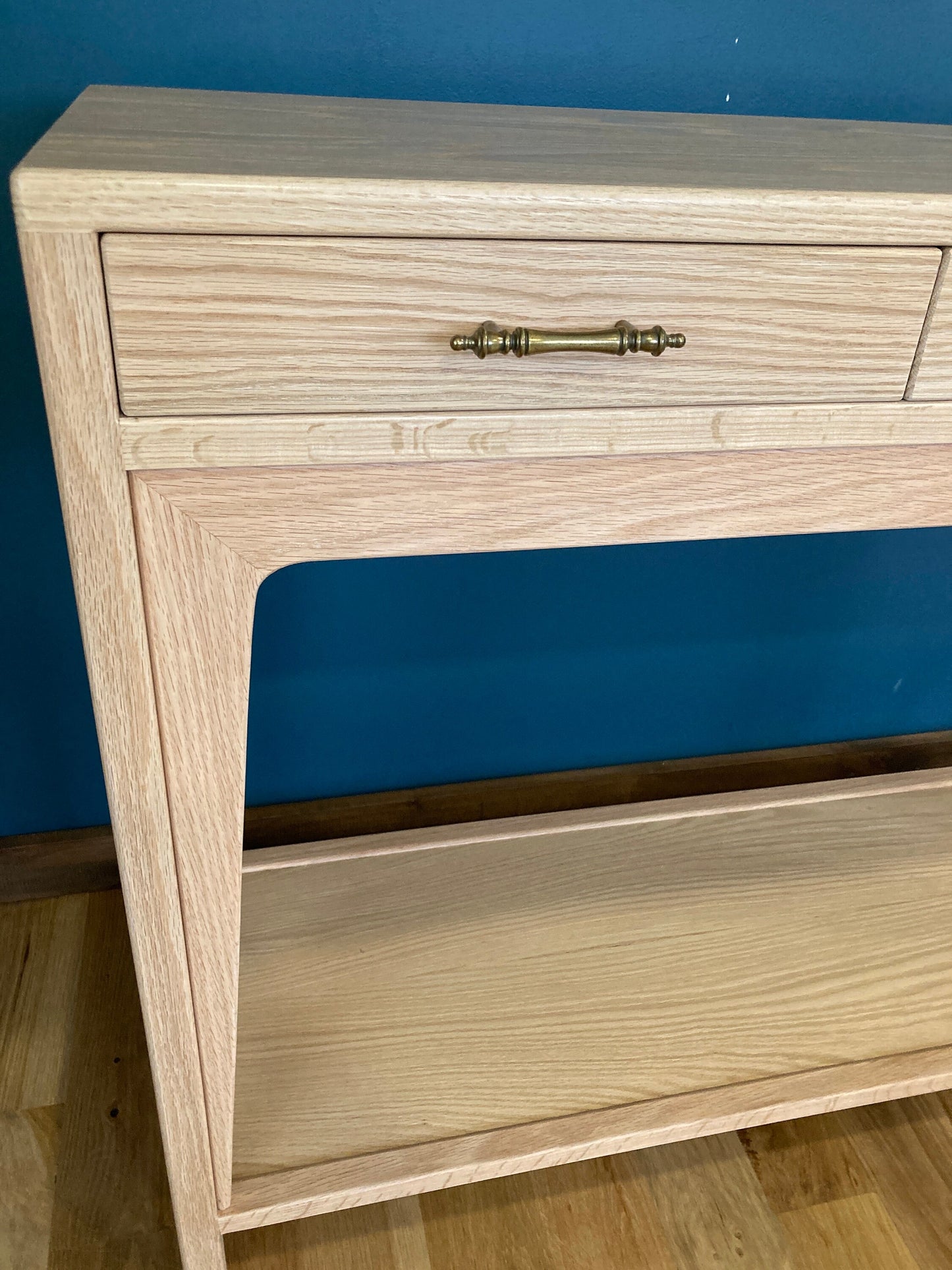 A light wood console table with a drawers and a lower shelf. The drawer has a brass handle. The table is on a wood floor.