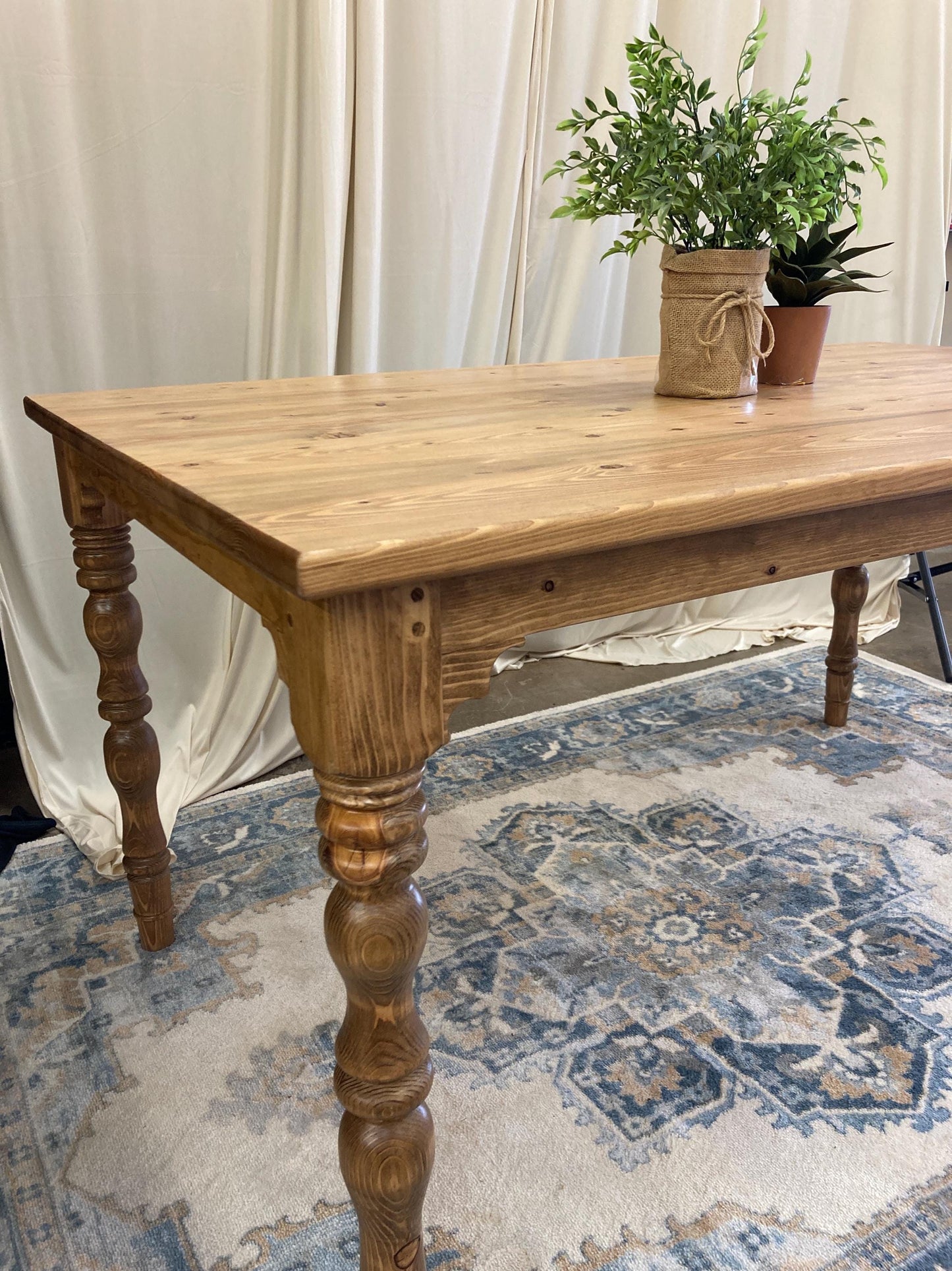 A wooden table with a light brown finish and turned legs. The table is on a rug with a blue and beige pattern.