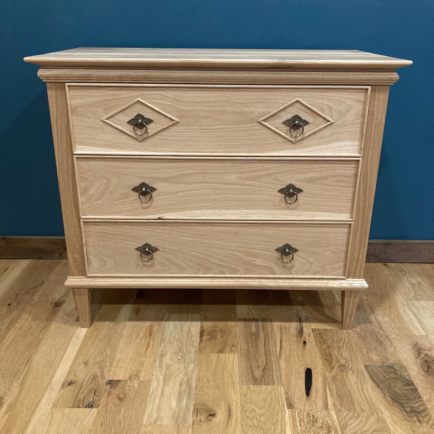 A light solid oak wood nightstand with three drawers and ornate metal handles. The dresser is in front of a blue wall and a wood floor.
