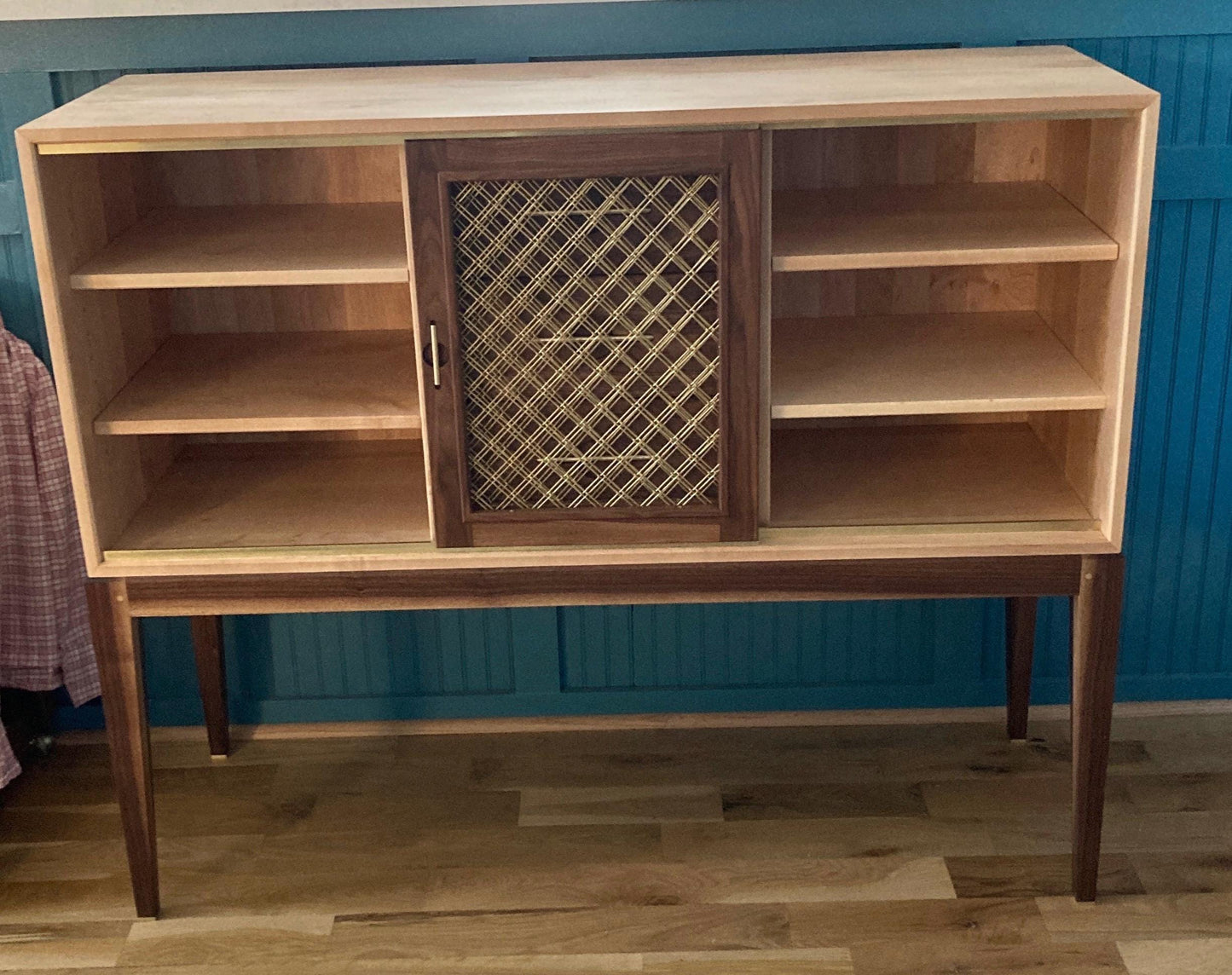 A wooden cabinet with two sliding doors. The cabinet has four shelves and is made of light wood with a dark wood trim. The sliding doors have brass mesh and is made of dark wood. The cabinet is standing on four legs.