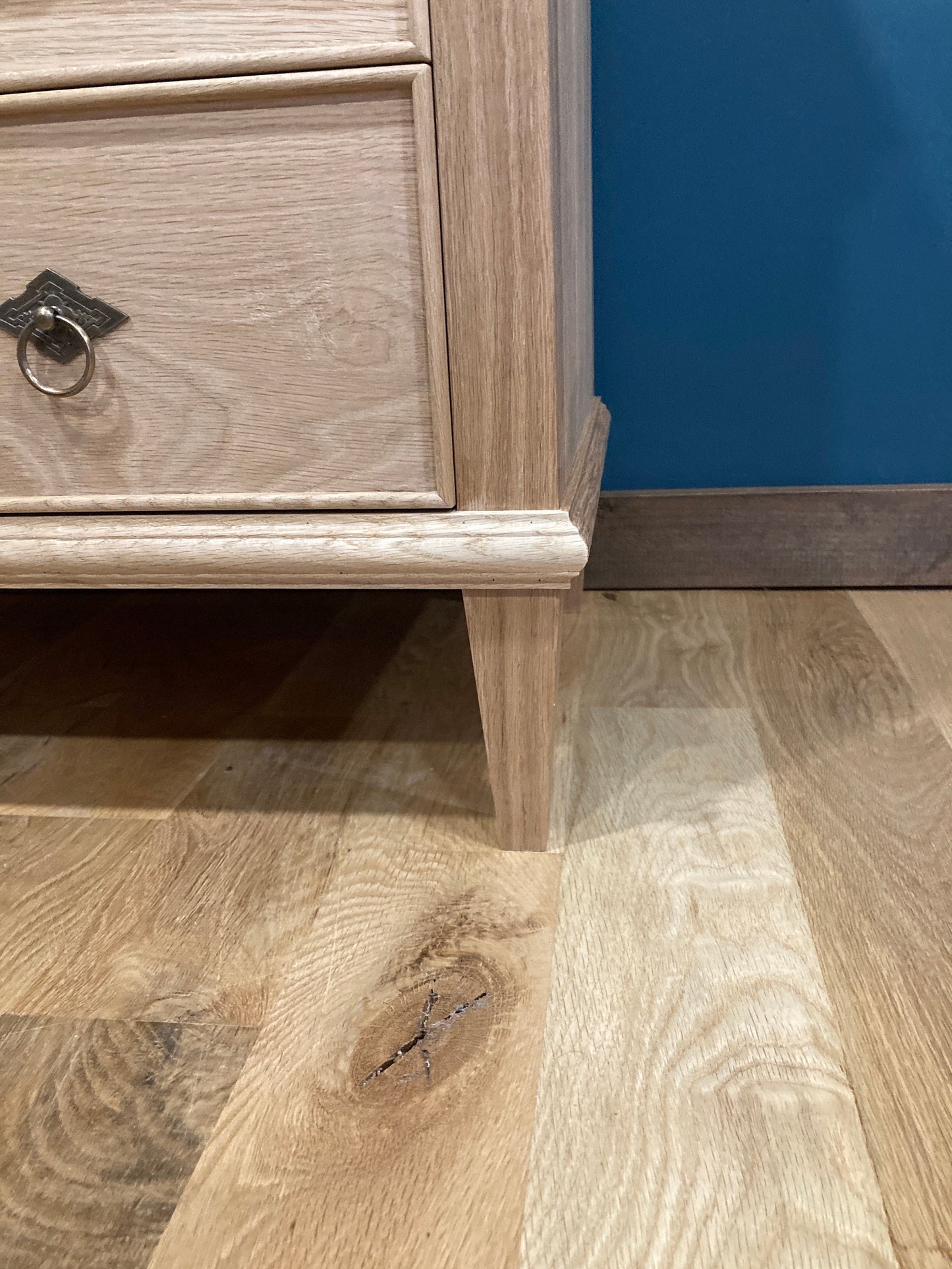 A light wood nightstand with a drawer and a decorative metal ring pull. The nightstand is on a light wood floor.