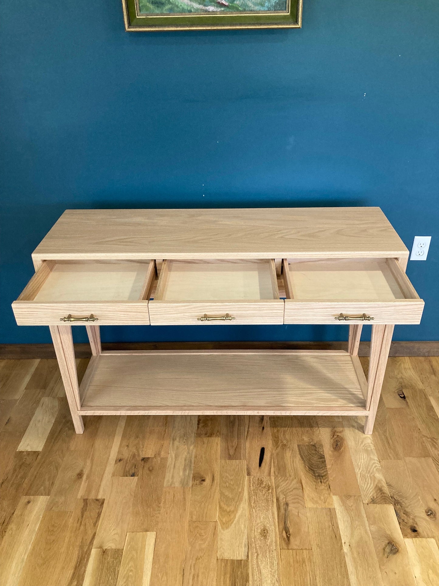 A light wood console table with three drawers and a lower shelf. The drawers are open and the table is on a light wood floor.