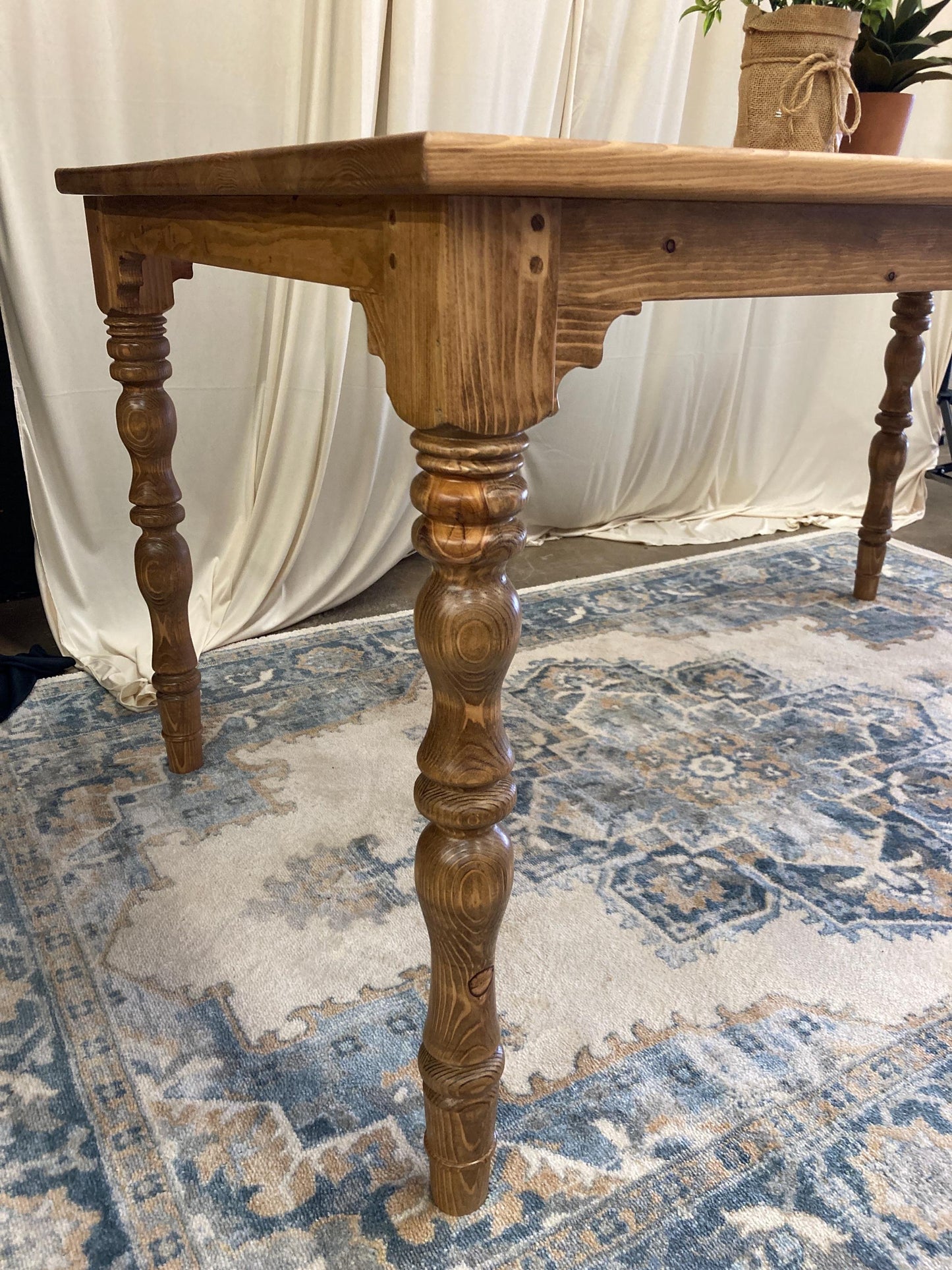 A close-up of a wooden table with turned legs. The table is made of light brown wood and has a simple, rustic design. The table is sitting on a patterned rug.