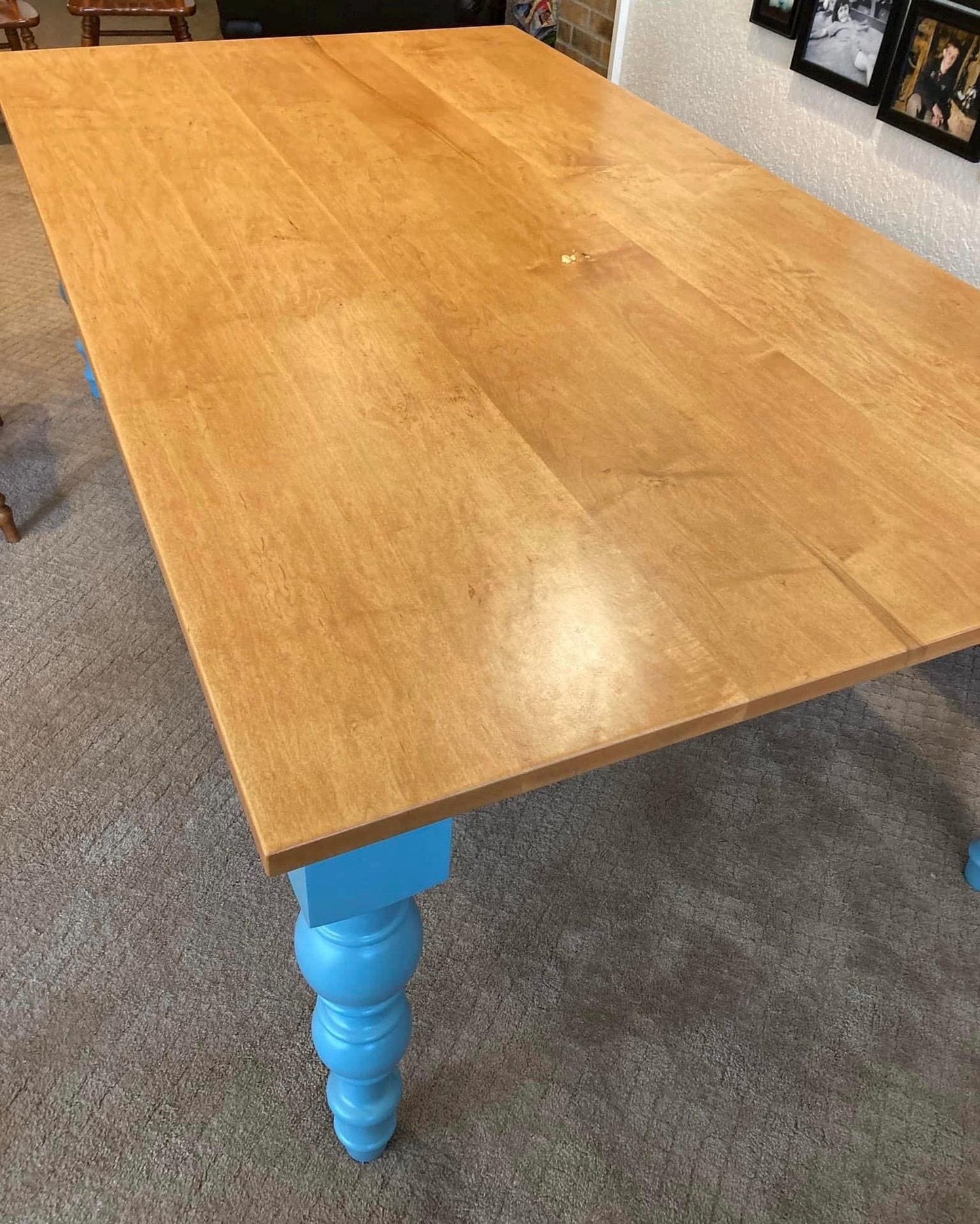 A light wood table with a blue painted base and hand turned legs. The table is in a room with a carpeted floor and a wall with framed pictures.