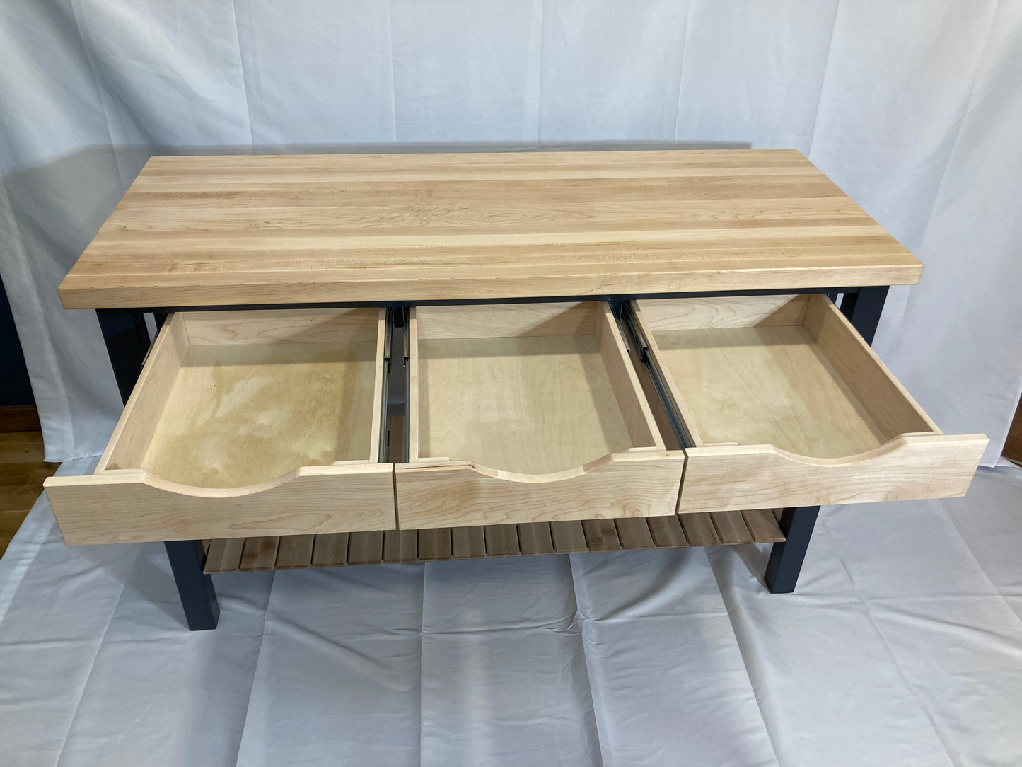 A kitchen island with a light maple butcher block top and three drawers. The drawers are made of light-colored wood and have a curved bottom. The island has a blue frame and a lower shelf. The drawers are open, showcasing the interior wood. This kitchen island is a functional and stylish addition to any kitchen.