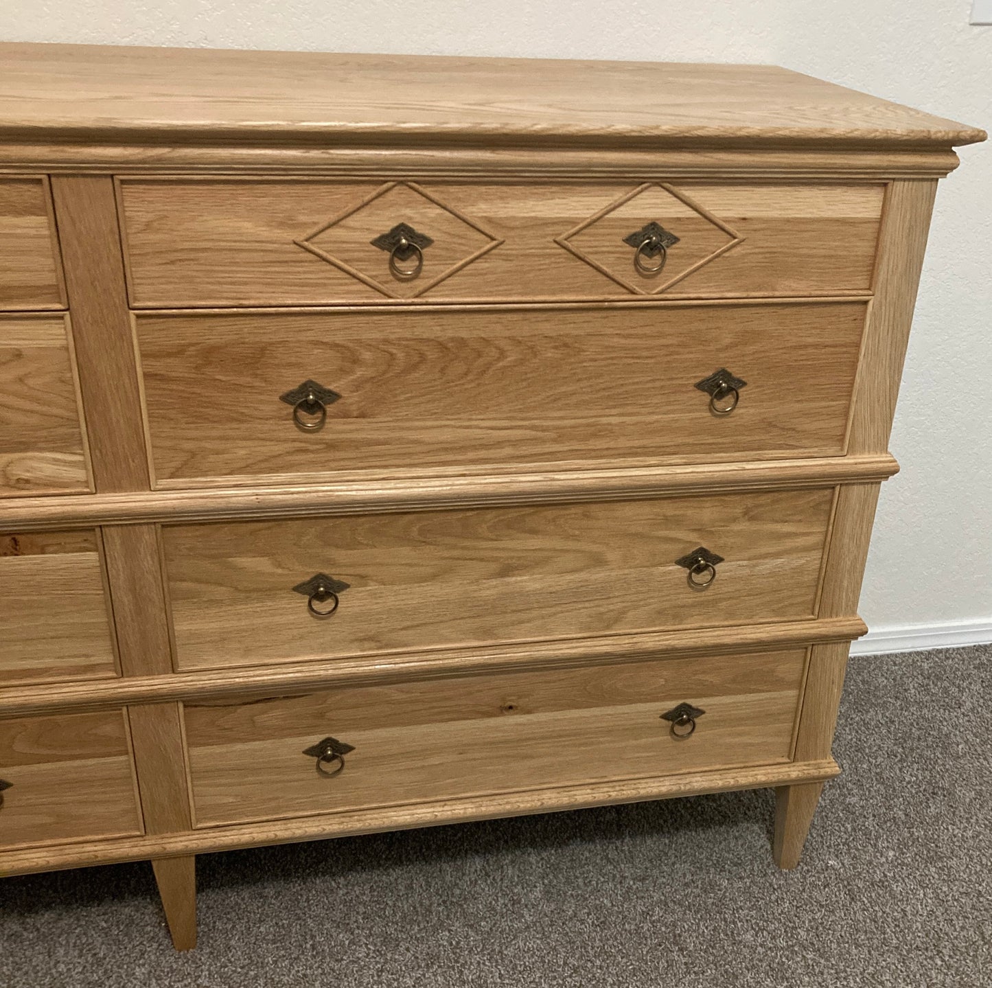 A light wood dresser with eight drawers and brass hardware. The dresser has a simple design with a diamond-shaped inlay on the top two drawers. The dresser is on a light brown carpet.
