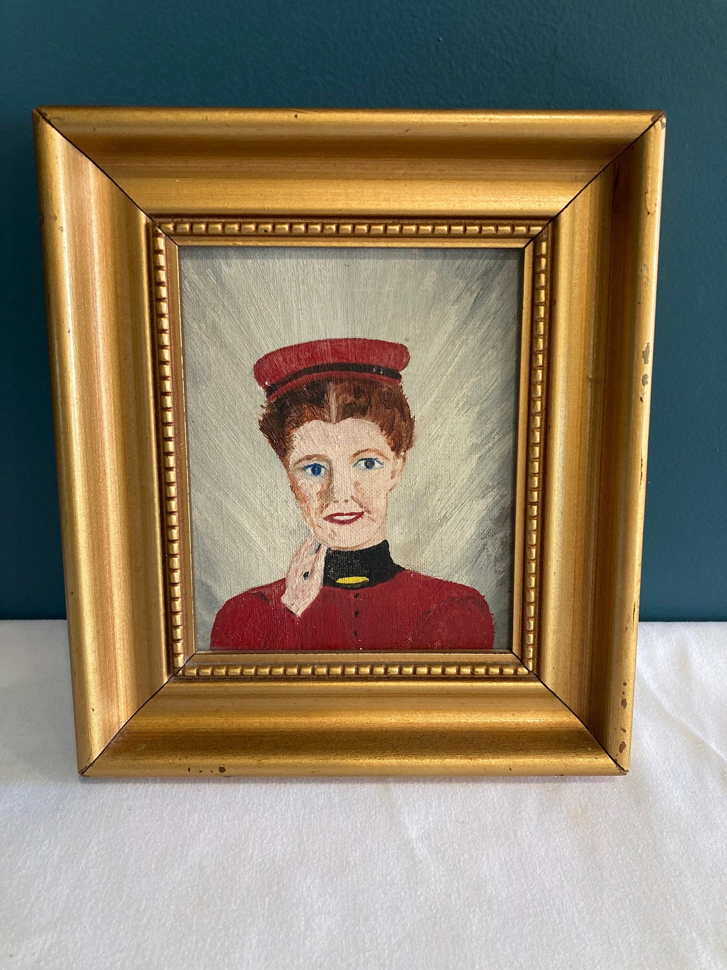 A framed painting of a woman wearing a red hat and a red dress with a black collar. The woman has her hand resting on the side of her face and is smiling and looking at the viewer.