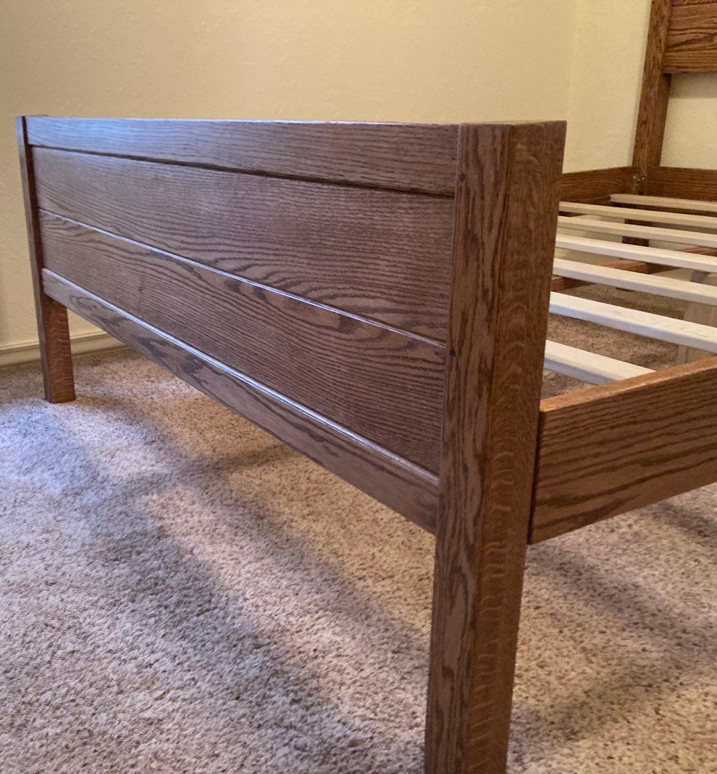 A wooden bed frame with a brown stain finish. The headboard is made of wood planks with a horizontal grain pattern. The footboard is also made of wood planks and has a similar grain pattern. The bed frame is sitting on a light brown carpet.