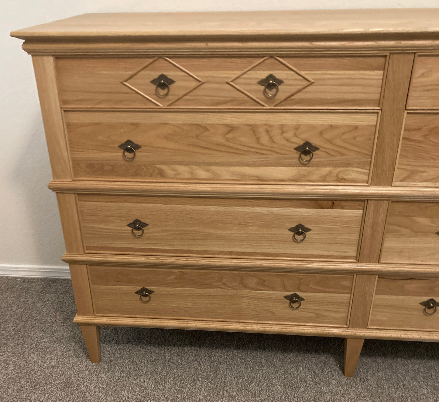 A light wood dresser with eight drawers and brass ring pulls. The top two drawers have a diamond-shaped design with a brass ring pull in the center. The dresser has a simple design with a light wood finish.