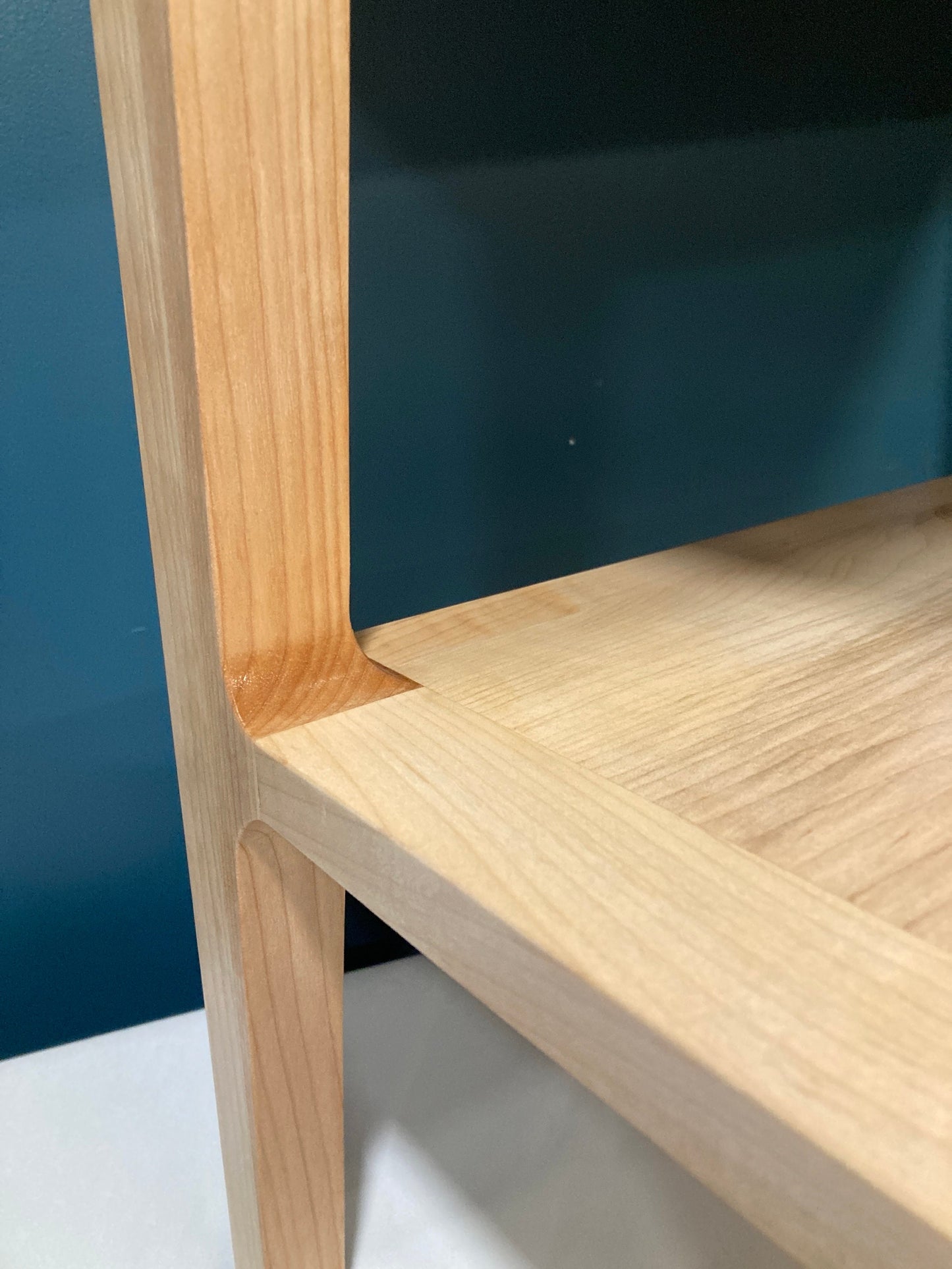 Close-up of a light natural wood side table with a minimalist design. The table features clean lines, a simple rectangular top, and sturdy legs. The wood grain is visible, showcasing the natural texture of the material. The joinery is precisely crafted, highlighting the quality of the woodworking. The light wood contrasts against an out-of-focus dark teal background.