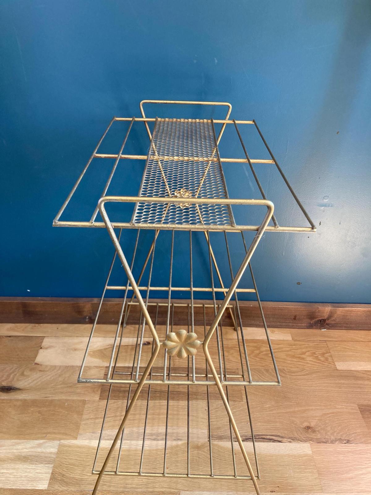 Gold metal three-tiered shelf with a geometric design. Each shelf is made of thin metal wires creating a grid pattern. The frame is gold-colored metal with decorative flower accents. The shelf is ideal for displaying items or storing small objects.