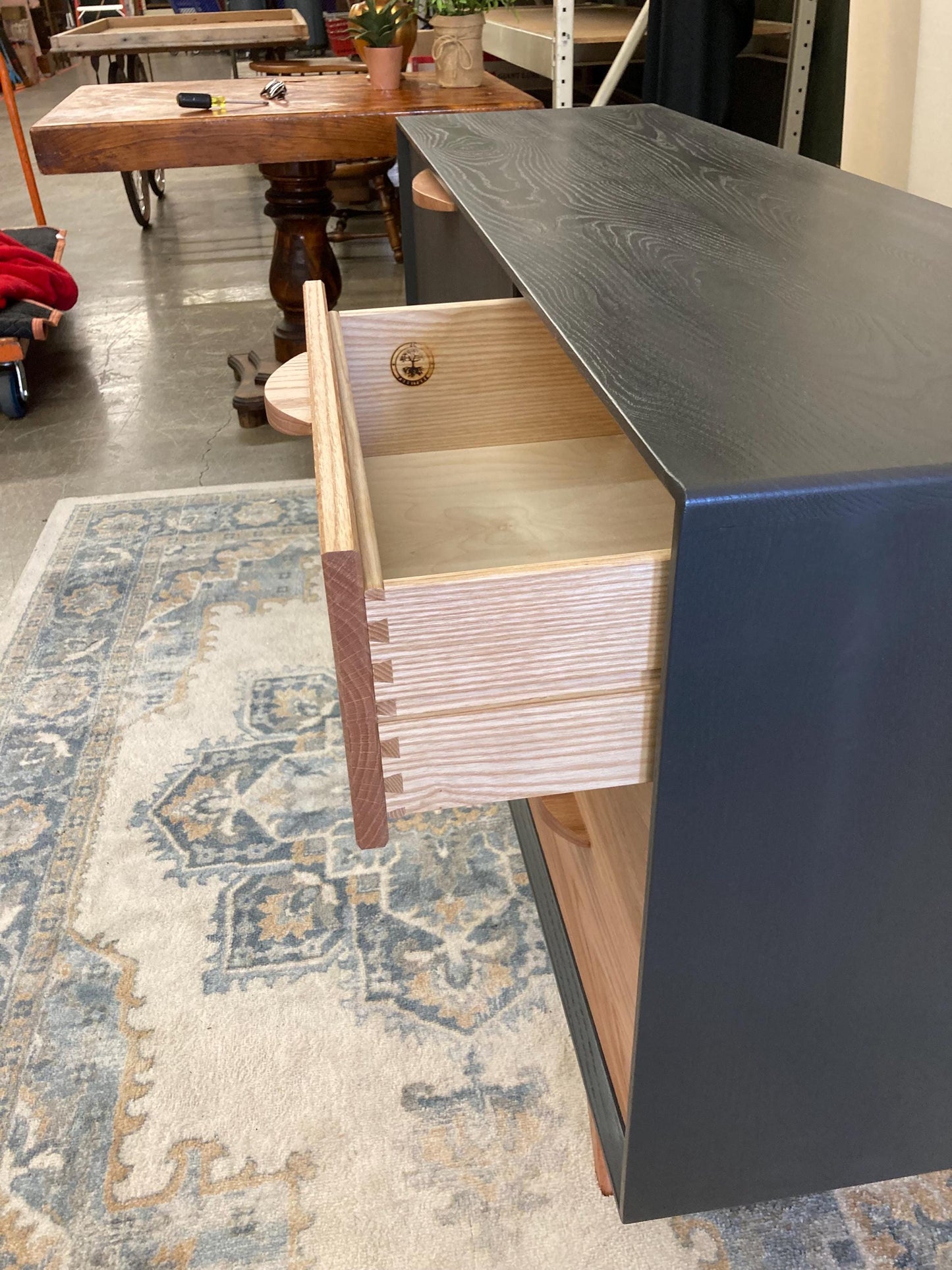 Close-up view of a open drawer. The drawer is light-colored wood with dovetail joinery visible. The dresser features a dark gray exterior finish. The drawer is partially open, revealing the interior wood. A small logo is visible inside the drawer. The dresser is positioned on a patterned rug.
