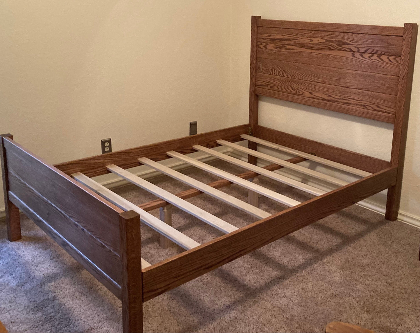 A wooden bed frame with a slatted headboard and footboard. The bed frame is made of dark brown wood and has a simple design. The bed frame is sitting on a light brown carpet.