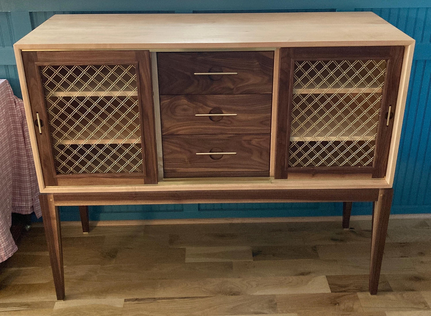 A wooden cabinet with three drawers and two brass mesh doors. The cabinet has a light wood top and dark wood sides and drawers. The doors have a brass mesh pattern. The cabinet is standing on four legs.