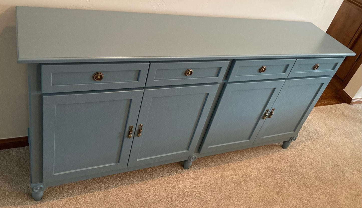 A light blue wooden cabinet with four doors and four drawers. The cabinet has brass knobs and is sitting on a light brown carpet.